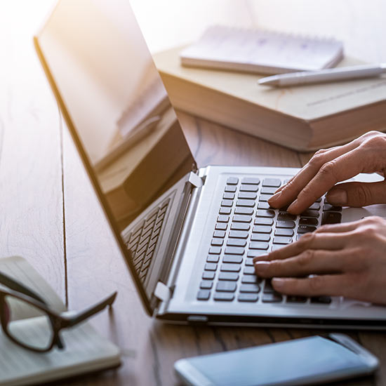 Winning-Local-Search Feature image. Person's hand resting on a laptop's keyboard while on a wooden desk with books, glasses, a phone, a notebook, and a pen around it.